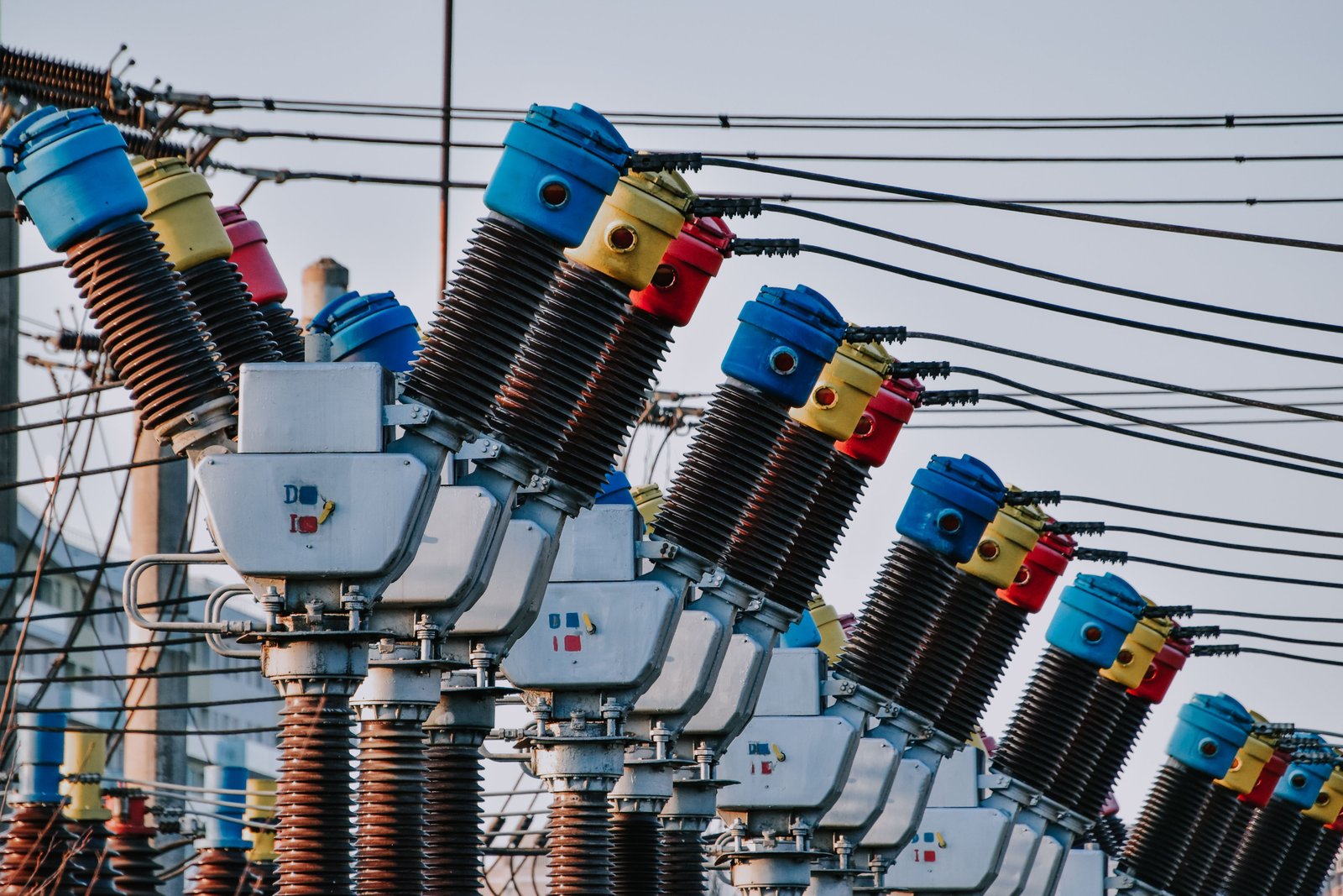 A row of high-voltage substation switchgear with red, yellow and blue insulator caps and copper windings against the sky.